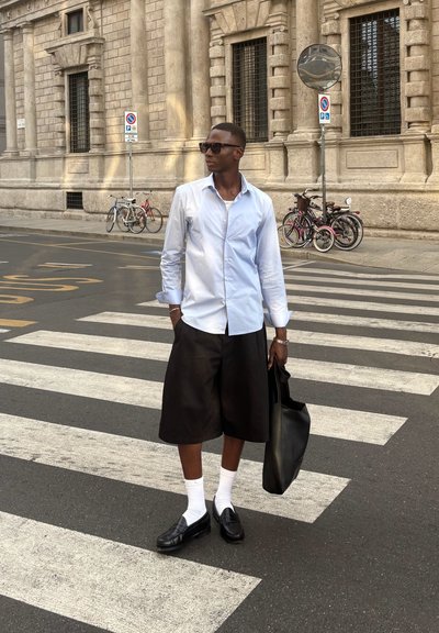 Camisa blanca de botones, pantalones cortos negros hasta la rodilla, zapatos negros con calcetines blancos, sosteniendo una bolsa negra, de pie en un paso de peatones con bicicletas de fondo.