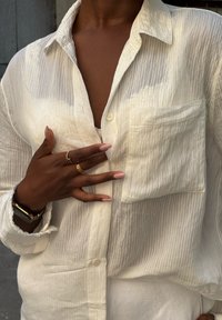 Camicia bianca a righe con bottoni e tasca sul petto. Tessuto stropicciato, colletto leggermente aperto. Mani decorate con anelli e un orologio.