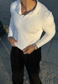 Person in einem weißen Langarm-Polohemd, schwarzen Hosen, silberner Uhr und Ringen, steht auf einem Steinpflaster im warmen Licht.