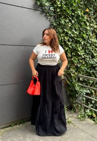 Hvid T-shirt med sort tekst og rød grafik, parret med en sort lagdelt maxi-nederdel og en lille rød taske. Grøn ivy i baggrunden.