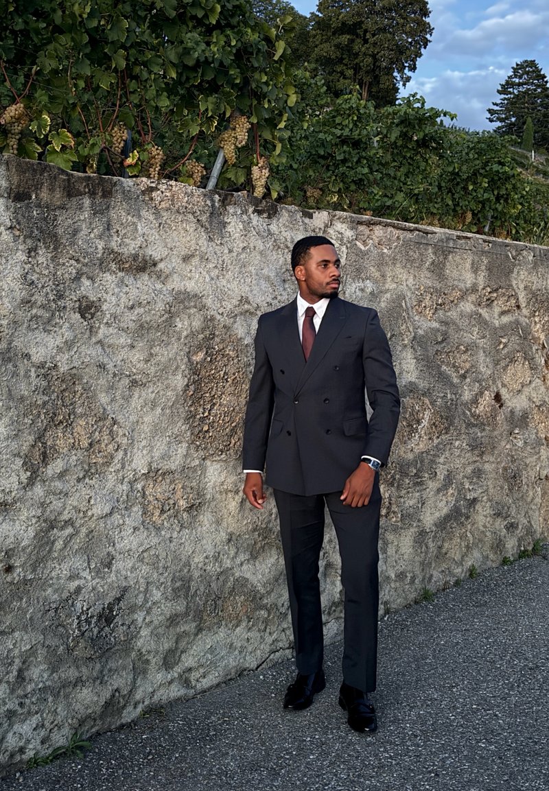 Homem em fato escuro de duplo peito e gravata castanha, em pé no passeio ao lado de um muro de pedra com folhagem verde e uvas por cima.