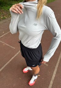 Osoba poprawiająca kołnierzyk jasnoszarej koszulki z długim rękawem, nosząca czarne szorty sportowe i białe-różowe buty na torze biegowym.