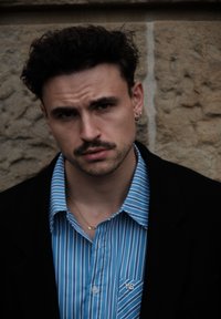 Mężczyzna z krótkimi, ciemnymi kręconymi włosami, ubrany w niebieską koszulę w paski z logo na kieszeni oraz czarną marynarkę, stoi na tle teksturowanej kamiennej ściany.