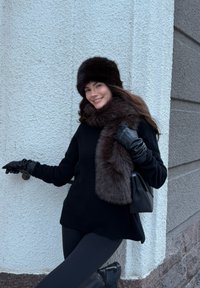 Femme en tenue d'hiver noire avec un chapeau en fourrure et une écharpe souriant, appuyée contre un mur gris clair texturé, tenant un sac noir et une rampe.