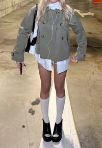 Oliivinvihreä lyhyt kaksirivinen bleiseri valkoisen paidan päällä; valkoiset polven yläpuolelle ulottuvat sukat ja mustat korotetut kengät. Sileät tekstuurit ja kontrastiset värit.