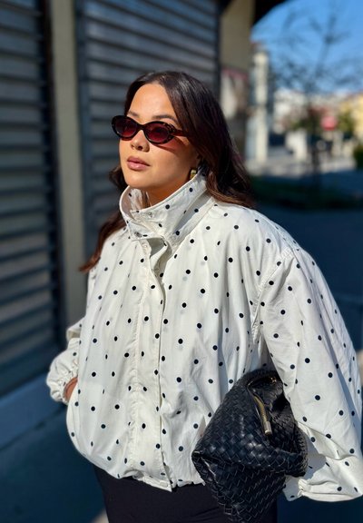 Mujer que lleva una chaqueta de lunares y gafas de sol con montura de ojo de gato, sosteniendo un bolso negro tejido y de pie al aire libre bajo la luz del sol.