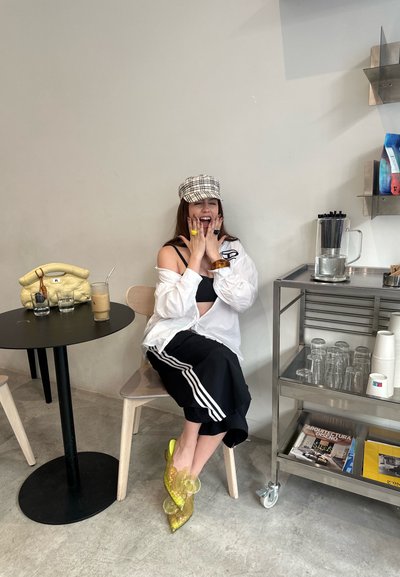Tacones de acrílico transparente con acentos florales, pantalones cortos negros con rayas blancas, camisa blanca oversize y una gorra a cuadros. Una bolsa amarilla cerca.