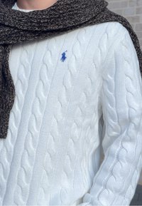 Pull blanc en maille torsadée avec un logo bleu brodé, accompagné d'une écharpe texturée brun foncé drapée autour du cou.