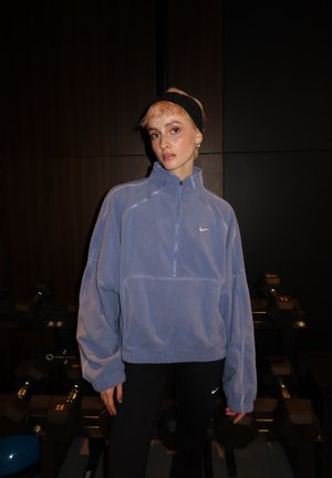 Joven con un forro polar azul de Nike y leggings negros, de pie en un gimnasio oscuro con pesas en el fondo.