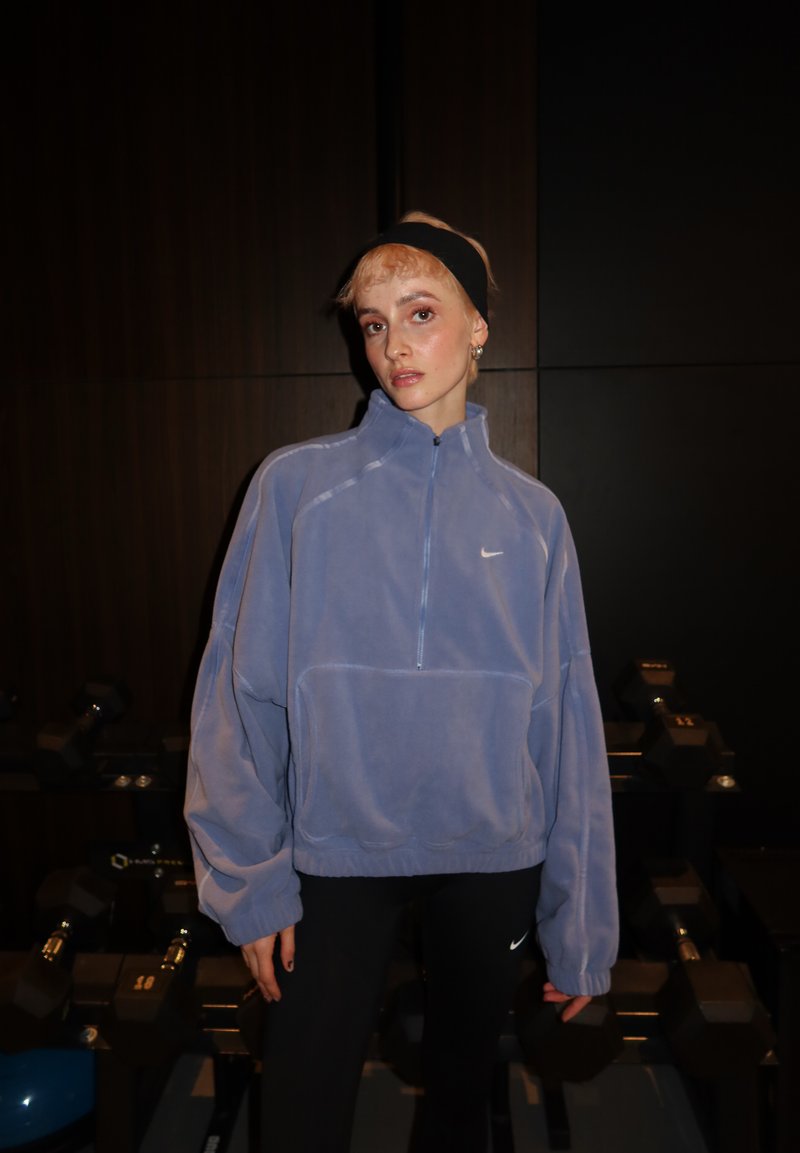 Jeune personne portant un sweat en polaire bleu Nike et des leggings noirs, debout dans une salle de sport tamisée avec des haltères en arrière-plan.