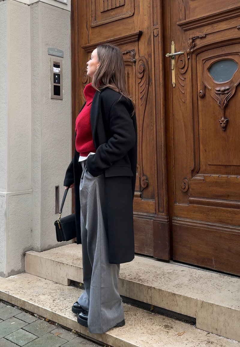 Donna con cappotto nero e maglione rosso in piedi su gradini di pietra, che tiene una borsa nera, guarda verso l'alto vicino a porte di legno ornate.