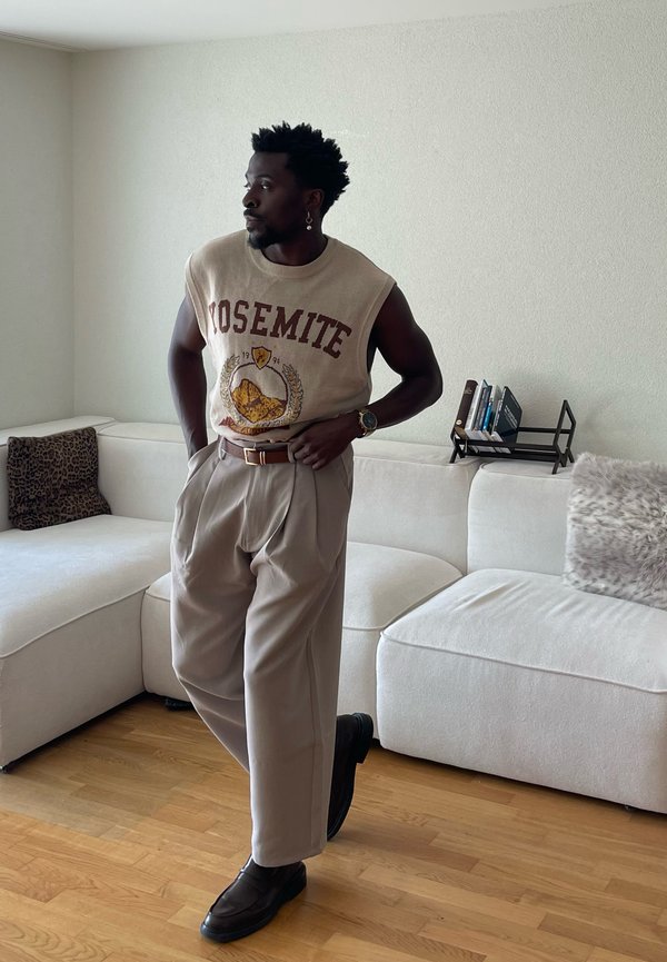 Mees, kes kannab beežikat varrukateta Yosemite kampsunit ja laiu pükse, seisab elutoas valge diivani ja puidust põrandaga.