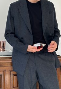 Person in dunkelgrauem Anzug und schwarzem Hemd hält eine geöffnete Parfümflasche und steht nahe einem Holzkabinett mit einer Kerze und einem Buch darauf.