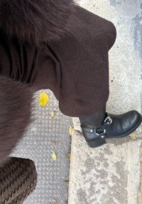 Ghete negre de piele până la gleznă cu o cataramă și o curea argintie, asortate cu un articol vestimentar texturat maro închis, pe o suprafață de pietriș cu frunze căzute.