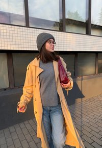 Trench bej, pulover gri și blugi albaștri. Ținând o geantă roșie lucioasă. Păr scurt, întunecat, cu o căciulă gri tricotată. Fundal urban.