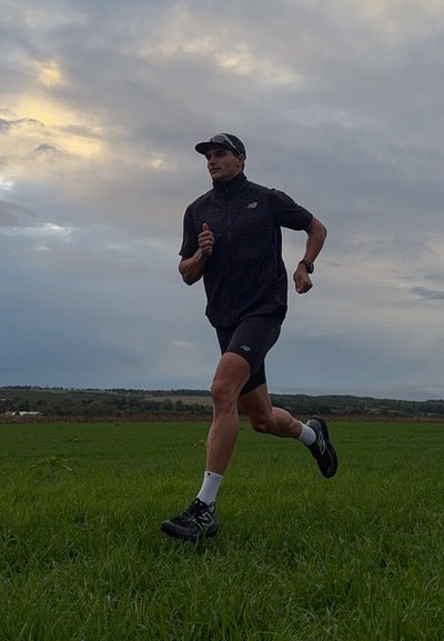 Chaqueta atlética negra y pantalones cortos, calcetines blancos y zapatillas de correr negras. El sujeto está corriendo sobre hierba verde bajo un cielo nublado.
