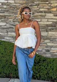 Λευκό strapless τοπ με συγκεντρωμένη μέση, συνδυασμένο με ανοιχτό μπλε τζιν. Τα αξεσουάρ περιλαμβάνουν χρυσά κοσμήματα και λευκά γυαλιά ηλίου. Φόντο από πέτρινο τοίχο.