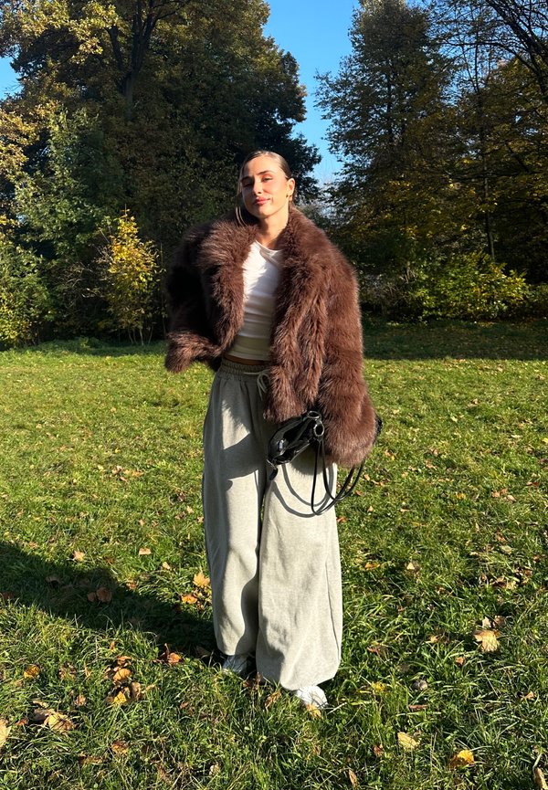 Brun faux fur jakke, hvid tætsiddende top, lys grå brede bukser og en sort håndtaske. Grøn græsbaggrund med efterårsblade.