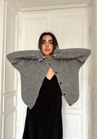 Cardigan gris surdimensionné en maille avec manches larges et fermeture boutonnée à l'avant, porté sur une robe noire, positionné devant un fond clair.