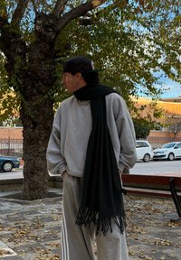 Sudadera gris de gran tamaño, bufanda negra con flecos, pantalones grises con rayas laterales, llevados con una gorra negra, de pie bajo un árbol con hojas de otoño.