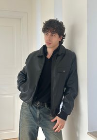 Zwarte leren bomberjack met een ritssluiting, geribde boorden en een borstzak, gecombineerd met een zwart shirt en vervaagde blauwe jeans.