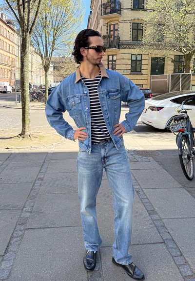 Hombre con gafas de sol que lleva chaqueta vaquera, camisa de rayas y vaqueros, de pie en una acera soleada de la ciudad con coches y bicicletas aparcados cerca.