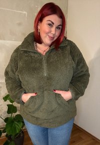 Sudadera de forro polar verde oliva con cuello de media cremallera, dos bolsillos frontales y una superficie suave y texturizada. Combinada con jeans azules.