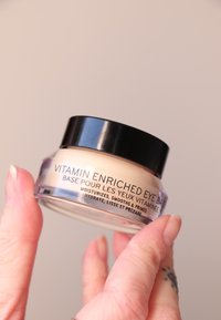 Vitamiinirikka silmäpohjustus kirkkaassa lasipurkissa, jossa on musta kansi. Sisällä näkyy kermainen beige tuote. Purkin tekstissä on tuotetiedot.