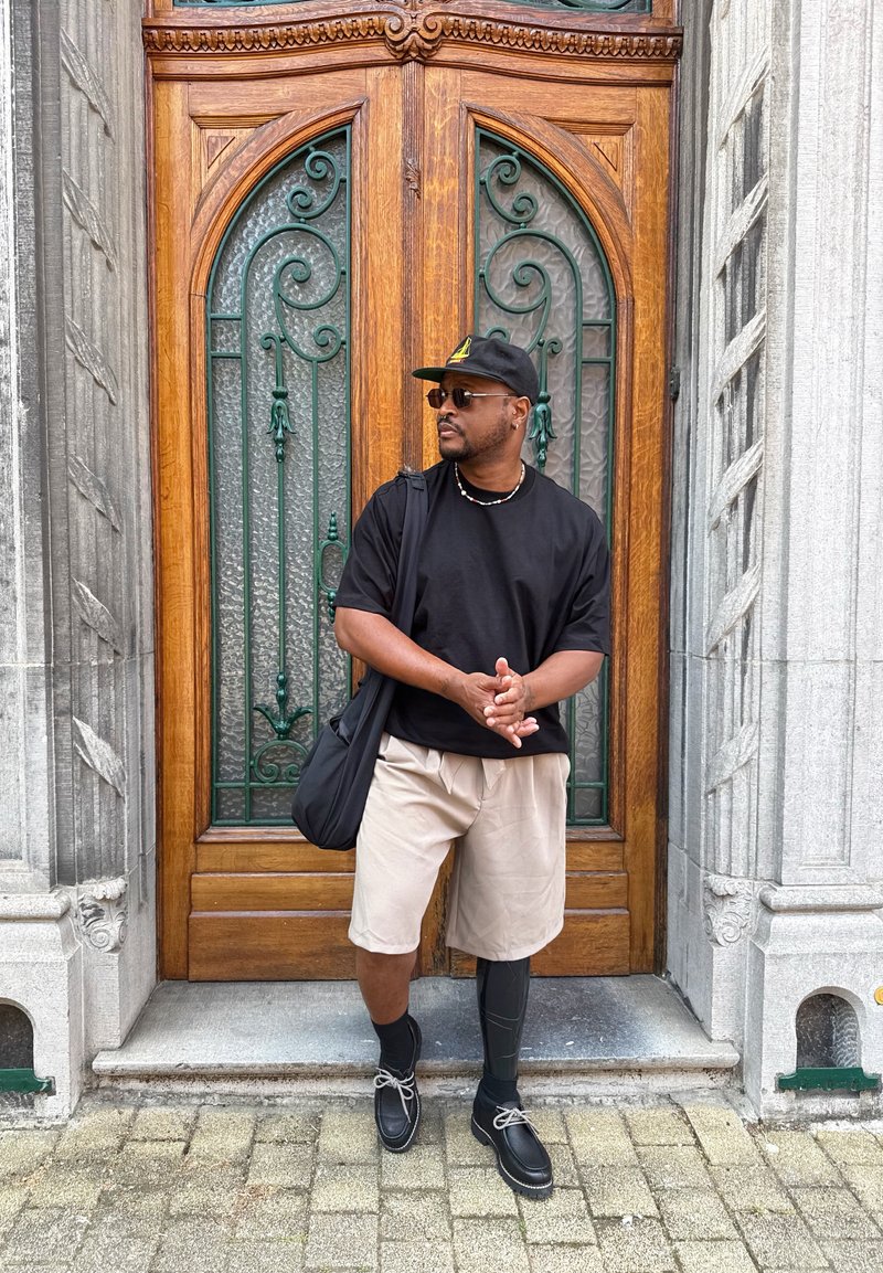 Homme avec une jambe prothétique portant une chemise noire, un short beige et une casquette, debout devant une porte en bois ornée de détails en fer forgé.