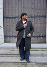 Manteau noir par-dessus un pull gris, pantalon foncé et baskets. Tient un sac bordeaux, posé près d'une porte en bois avec des ferrures décoratives.