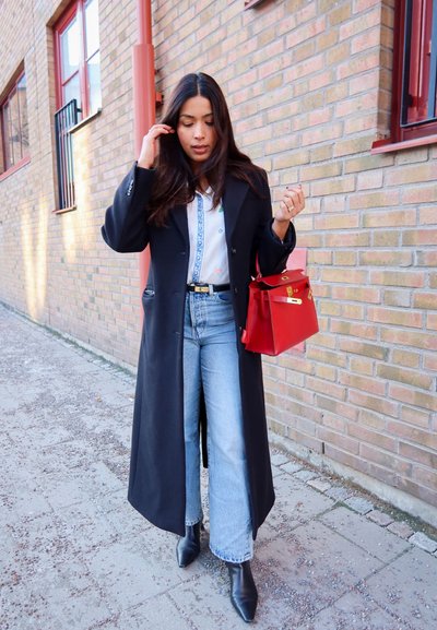 Mujer con cabello largo y oscuro, vestida con abrigo negro, camisa blanca estampada, jeans azules y botas negras, sosteniendo un bolso rojo mientras camina por una acera cerca de una pared de ladrillos.