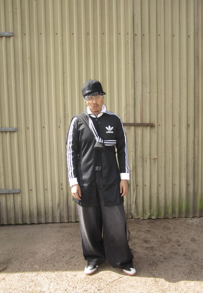 Veste noire zippée avec des rayures blanches, associée à un pantalon en denim foncé ample. Les accessoires incluent une casquette noire et des lunettes de soleil, debout sur une surface en terre.