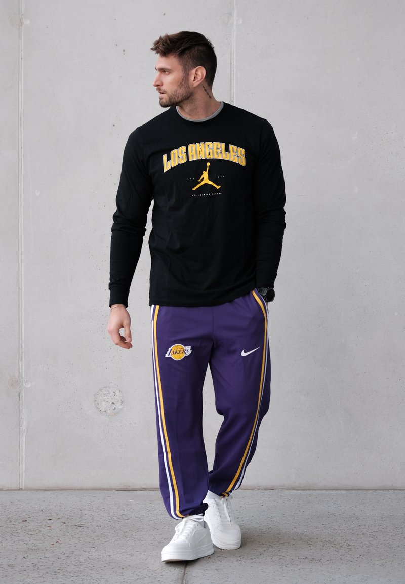 Chemise noire à manches longues avec "LOS ANGELES" et un logo jaune, associée à un pantalon de survêtement violet avec des bandes jaunes et un logo des Lakers, baskets blanches.