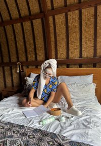 Persoon in een blauw bloesemshirt, met een handdoek op het hoofd, schrijft in een notitieboek op een bed met witte lakens, omringd door skincareproducten en voedsel.