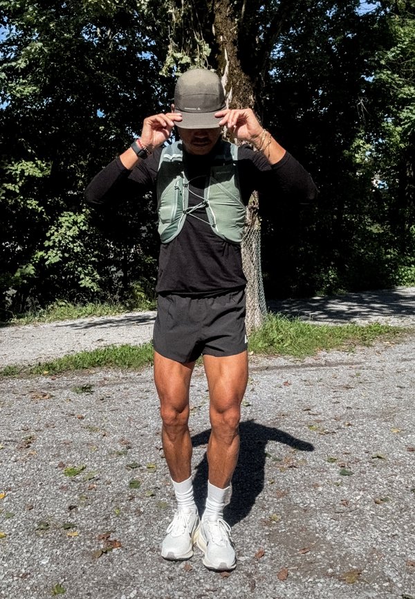 Person udendørs justerer en kasket, iført løbevest, sort trøje, shorts, hvide sokker og sneakers, stående på en grussti.