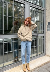 Fiatal vörös hajú személy bézs kapucnis pulóverben, világoskék farmerban és világosbarna csizmában áll egy nagy üvegajtónál, oldalra néz, kezét a fejére téve.