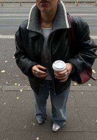 Μαύρο δερμάτινο μπουφάν με γκρι ριγέ γιακά, συνδυασμένο με ανοιχτό γαλάζιο φαρδύ παντ�ελόνι και γκρι αθλητικά παπούτσια. Κρατάει ένα φλιτζάνι καφέ και μια μπορντό τσάντα.