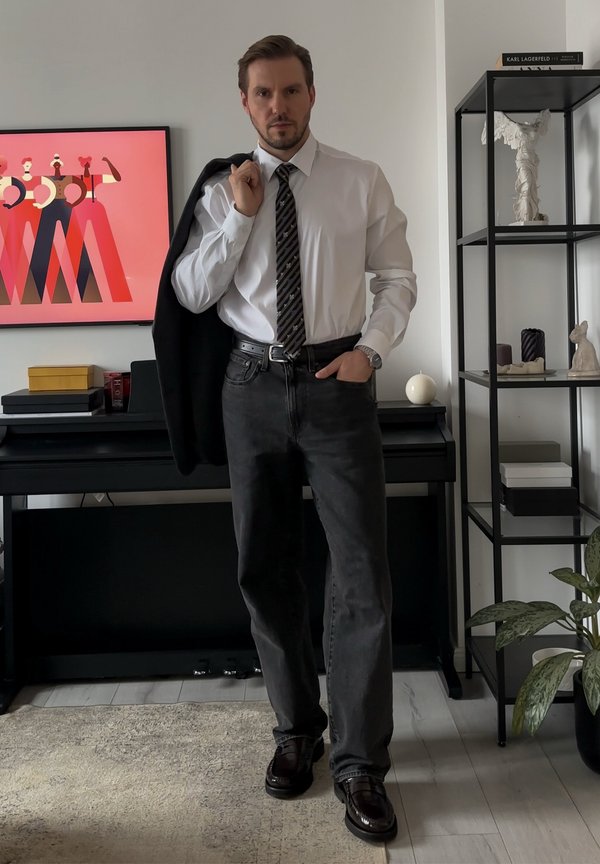 Mees valges särgis, triibulise lipsu ja tumedate teksadega hoiab musta jopet õlal moodsas elutoas, kus on kunst ja riiulid.