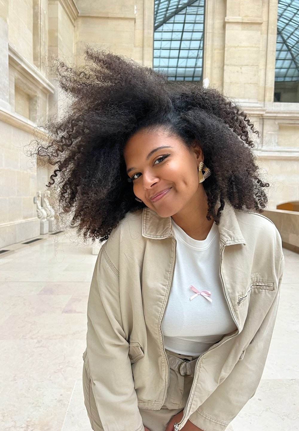 Veste beige zippée portée sur une chemise blanche avec un détail en ruban rose. Le mannequin a des cheveux foncés, volumineux et bouclés, ainsi que des boucles d'oreilles dorées. Cadre naturel.