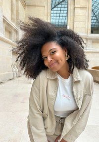 Veste beige zippée portée sur une chemise blanche avec un détail en ruban rose. Le mannequin a des cheveux foncés, volumineux et bouclés, ainsi que des boucles d'oreilles dorées. Cadre naturel.