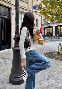 Vrouw in trui en jeans stelt sneaker bij terwijl ze koffie en bloemen vasthoudt op een stadsstoep nabij een lantaarnpaal en een herfstboom.