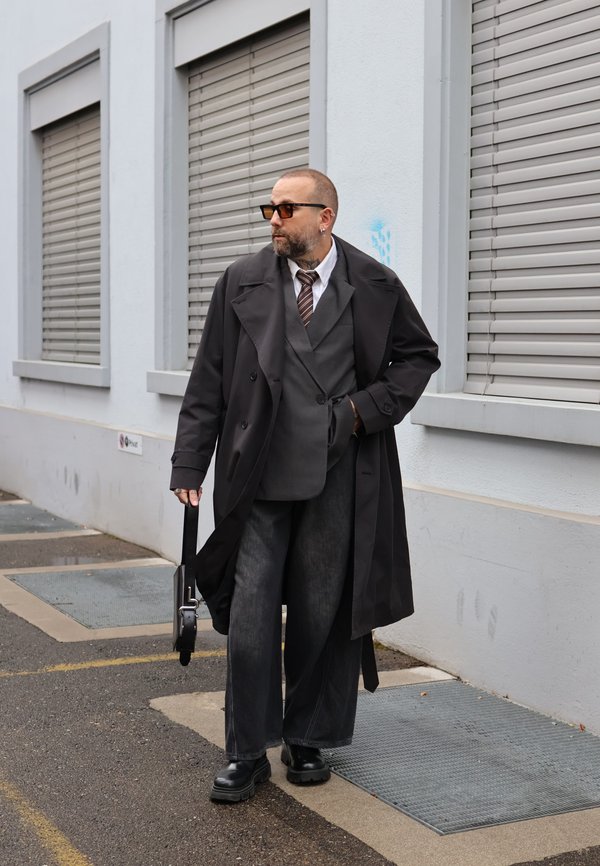 Un hombre con barba, gafas de sol y un abrigo largo negro está de pie junto a un edificio y sostiene una pequeña maleta negra.