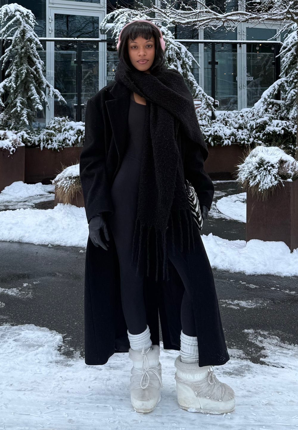 Long manteau noir, écharpe noire et gants, assortis à un body noir et des bottes blanches doublées de fourrure. Fond enneigé avec des arbres à feuilles persistantes.