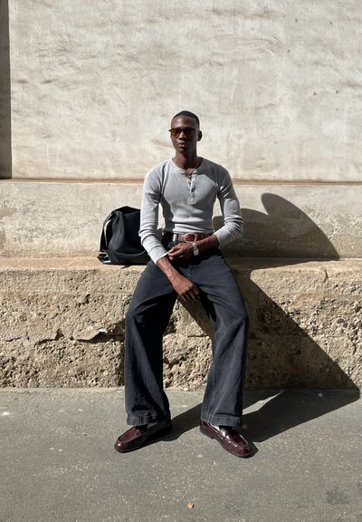 Camisa henley de manga larga gris combinada con pantalones de pierna ancha negros y mocasines burdeos. Un reloj de correa de cuero y gafas de sol completan el look.