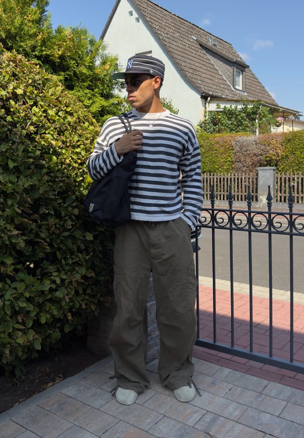 Homme portant une chemise rayée à manches longues, un pantalon kaki et une casquette se tient près d'une cloture métallique près de buissons verts et d'une maison par temps ensoleillé.