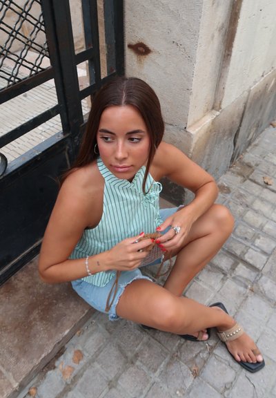 Top halter de rayas en verde claro con detalle de lazo, combinado con shorts de denim desgastados. Presenta sandalias planas con tachuelas y un bolso de mimbre.
