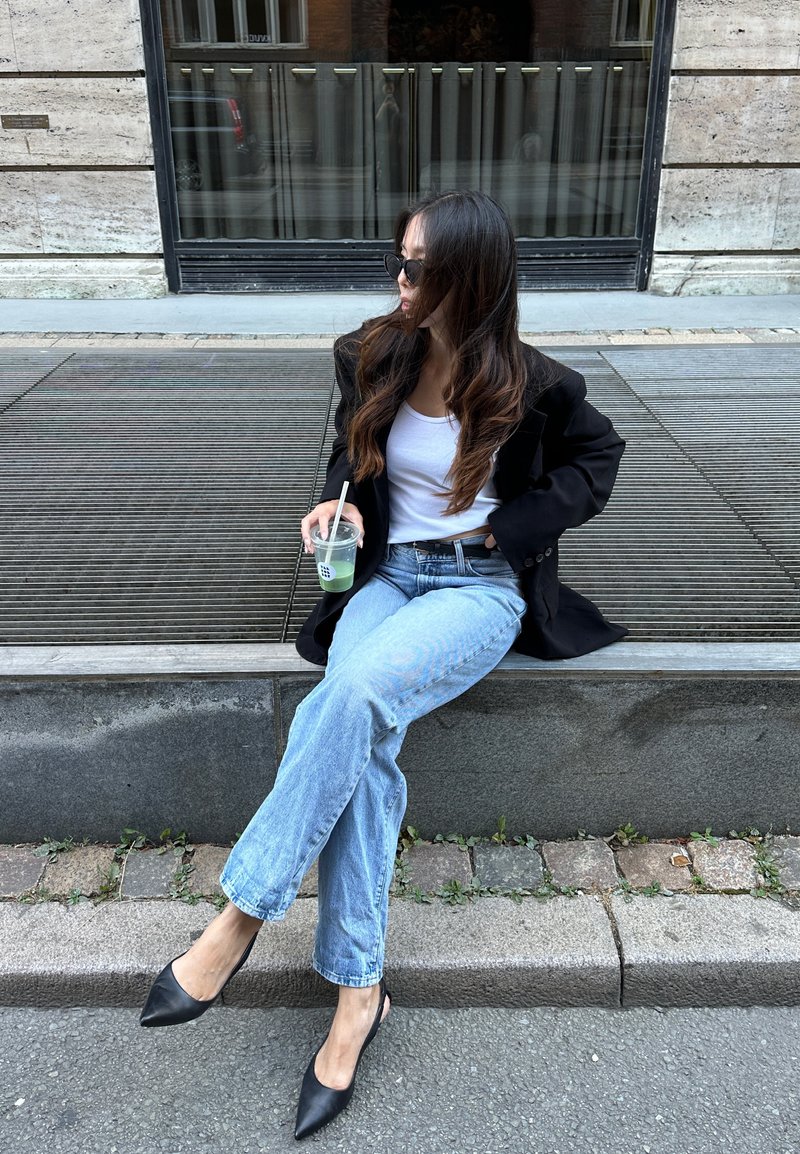Czarna, oversize'owa marynarka, biała bluzka na ramiączkach, jasnoniebieskie dżinsy i czarne, spiczaste buty na obcasie typu slingback. Trzyma przezroczysty kubek z zielonym napojem.