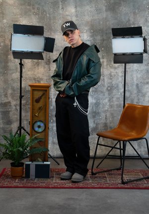 Joven con gorra negra y chaqueta verde, de pie entre dos luces de estudio, junto a una silla marrón, un mueble vintage, una radio y una planta en maceta.