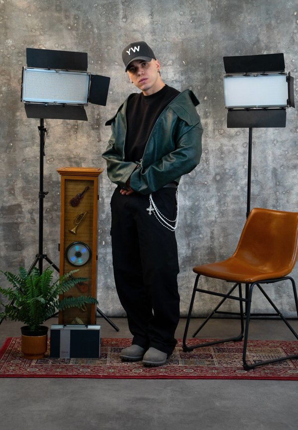 Young man wearing a black cap and green jacket stands between two studio lamps, beside a brown chair, a vintage cabinet, a radio, and a potted plant.