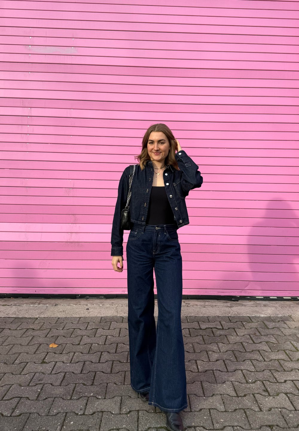 Temno modra jeans jakna in široke kavbojke z visokim pasom, združene s črnim topom. Dodatki vključujejo večplastne ogrlice in majhno torbico. Rožnata ozadje.